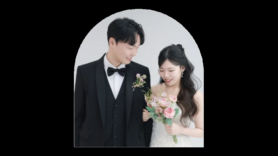 턱시도를 입은 남편과 꽃다발을 든 아내가 서로 바라보며 웃는 웨딩 사진