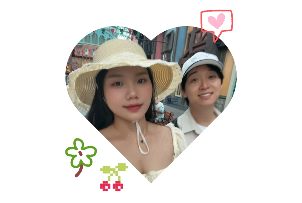 알록달록한 건물이 있는 거리에서 밀짚모자를 쓴 아내와 모자를 쓴 남편이 함께 웃는 하트 모양 사진