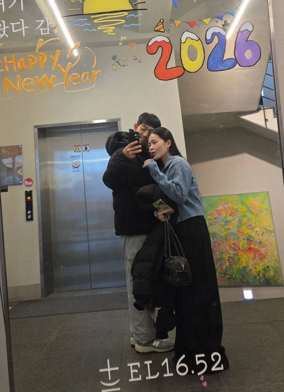 2026 Happy New Year 그림이 그려진 엘리베이터 앞 거울에서 함께 셀카를 찍는 부부