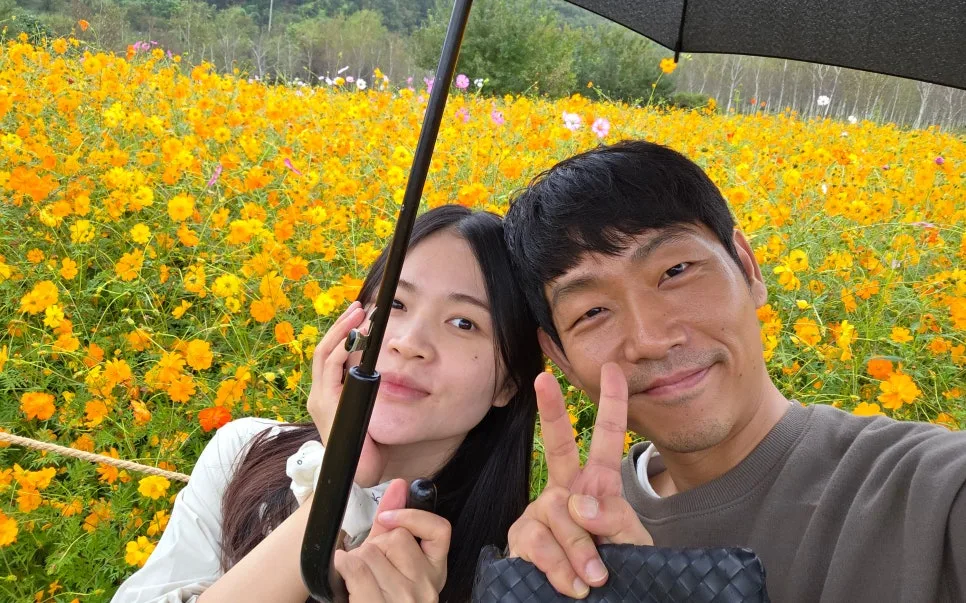 노란 코스모스 꽃밭에서 우산을 함께 쓰고 셀카를 찍는 윤현준 님 부부