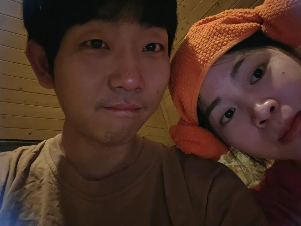 나무 벽으로 된 방에서 주황색 수건 모자를 쓴 아내분과 남편이 함께 찍은 셀카