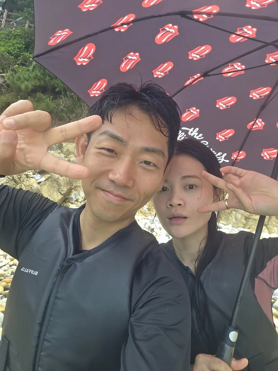 자갈 해변에서 비를 피해 우산을 함께 쓰고 브이 포즈를 하는 부부, 아내분 손에 반지가 보임